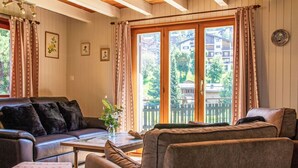 Ein gemütliches Wohnzimmer mit Sofa, Sesseln und Couchtisch. Vorhanden sind Vorhänge, gerahmte Bilder an der Wand und ein Blick auf Gebäude durch große Glasüren.
