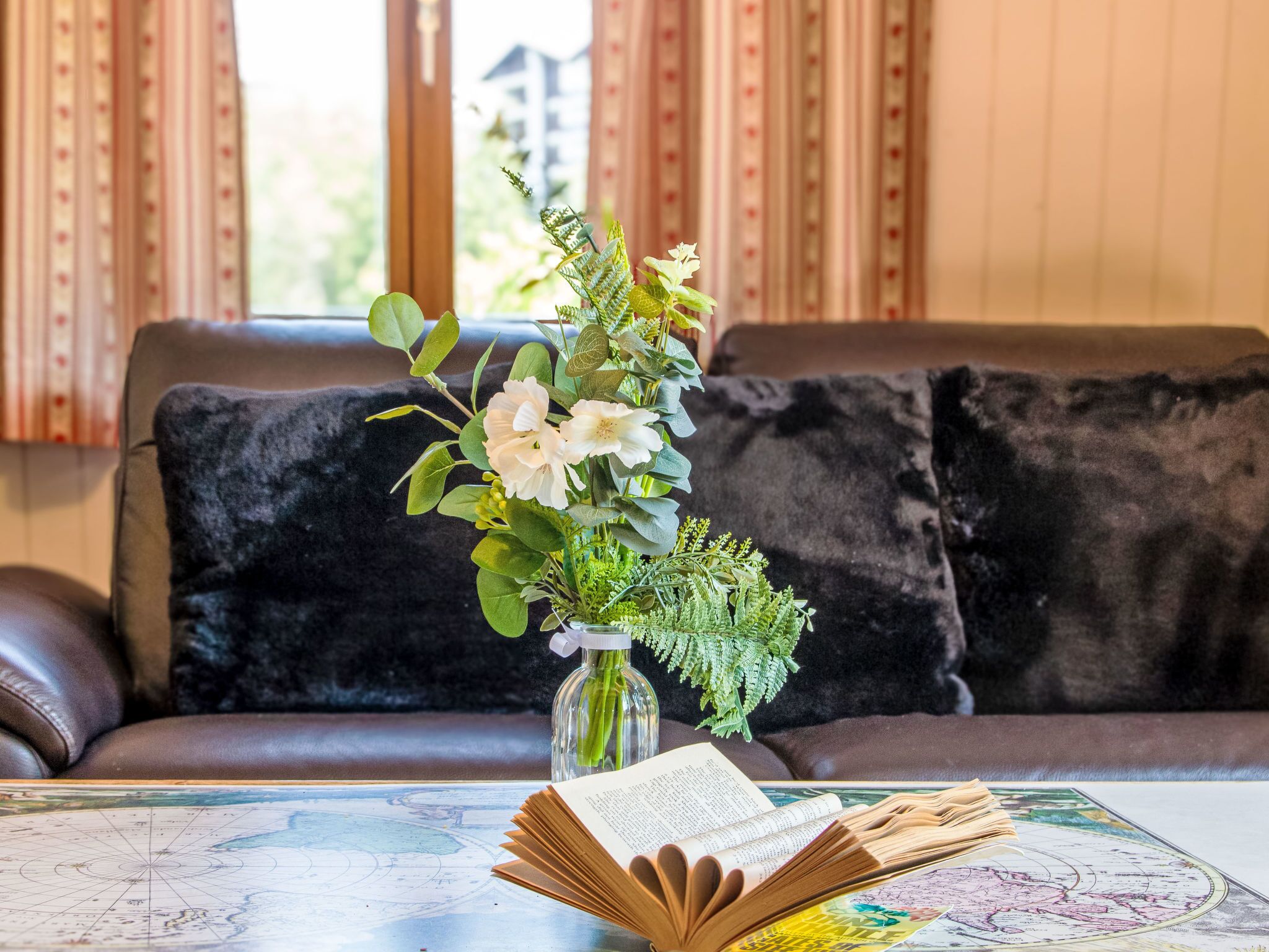 Une pièce avec un canapé en cuir, un coussin noir, un vase de fleurs, un livre et une carte sur la table.
