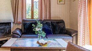 Ein gemütliches Wohnzimmer mit einem Ledersofa, einem Couchtisch mit Büchern und einer Blumenvase sowie einem Fenster mit Vorhängen.