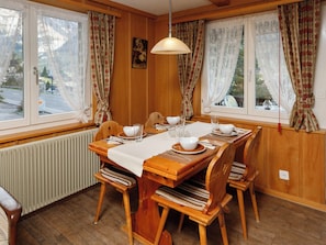 A wooden dining room with a table set for four, large windows with curtains, a radiator, and a hanging light fixture.