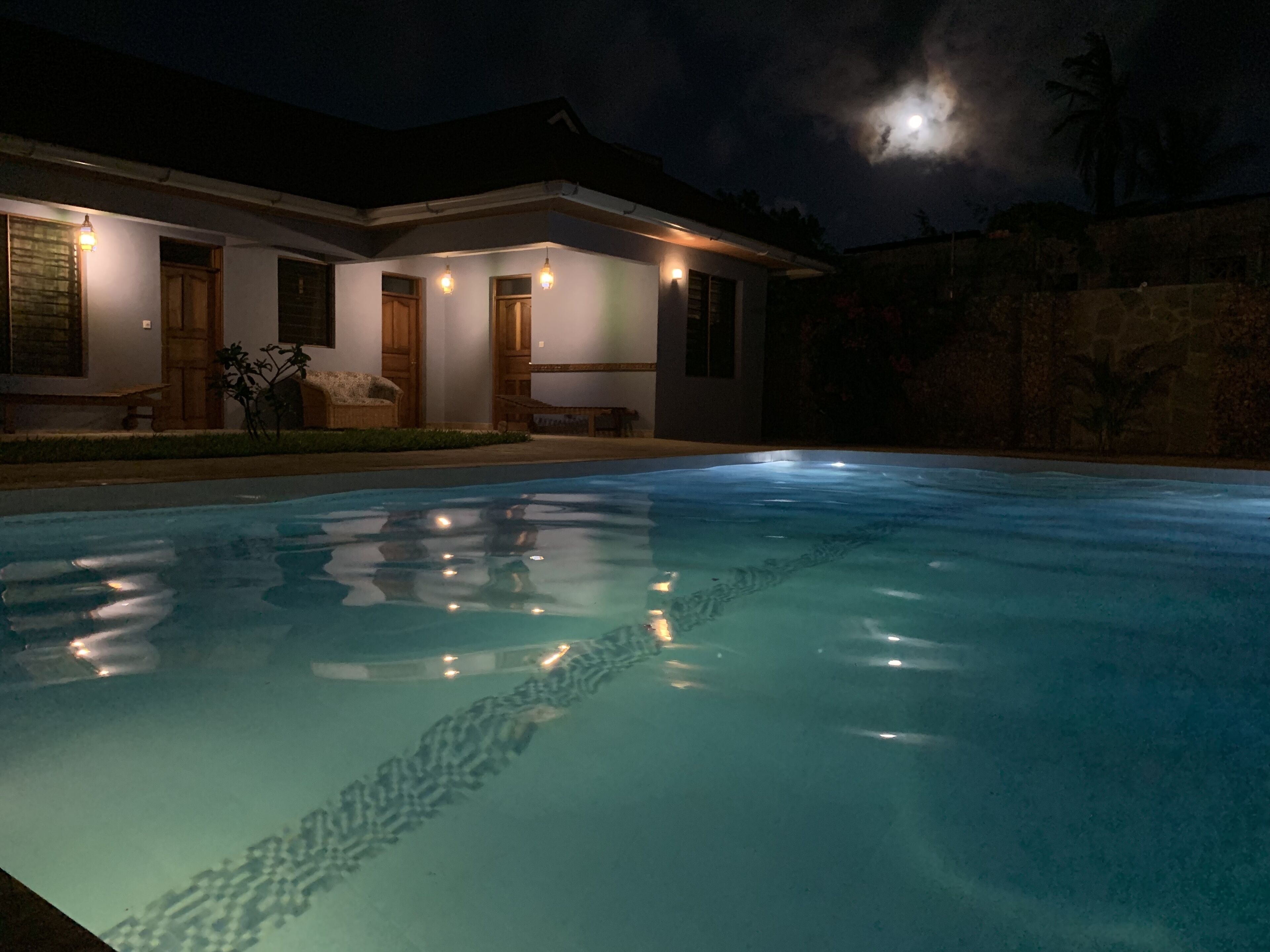 A swimming pool with a clear water surface reflecting lights, surrounded by a building with multiple doors and windows, and a night sky with a visible moon.