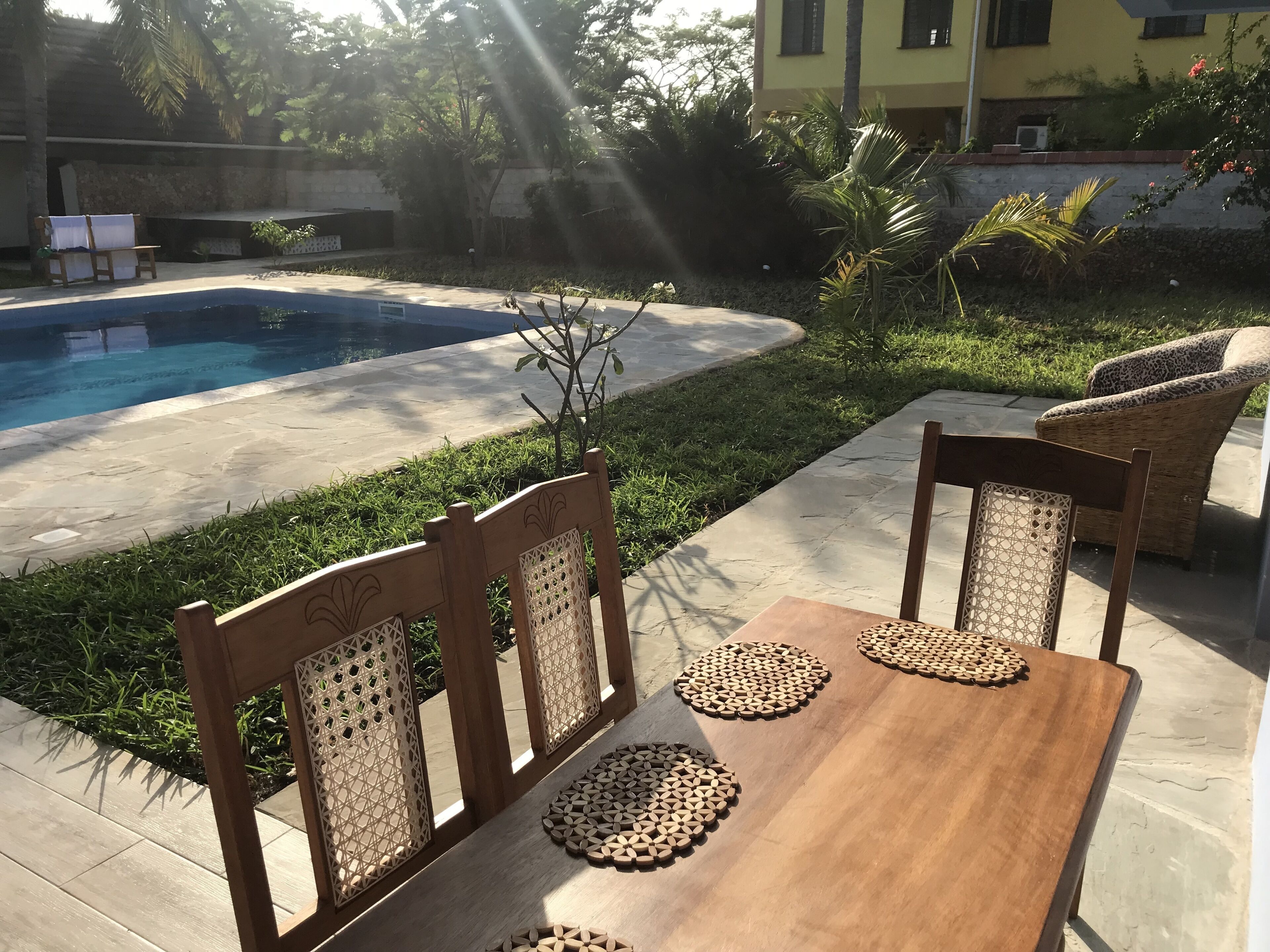 Una mesa de madera con individuales tejidos y sillas de mimbre, ubicada en un patio con piscina al fondo.