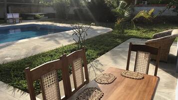 Una mesa de madera con individuales tejidos y sillas de mimbre, ubicada en un patio con piscina al fondo.