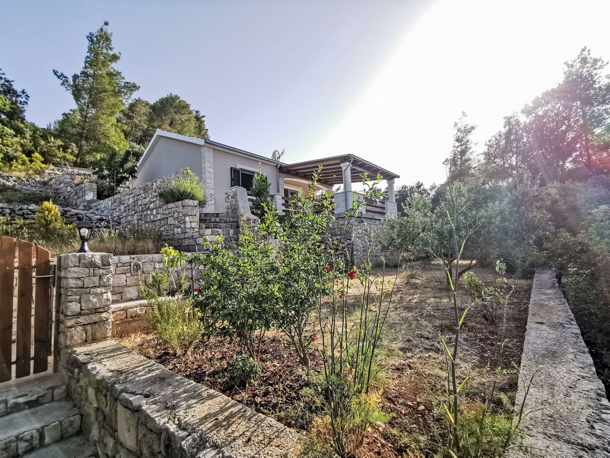 Une maison avec un mur en pierre, un portail en bois et un jardin où poussent différentes espèces de plantes.