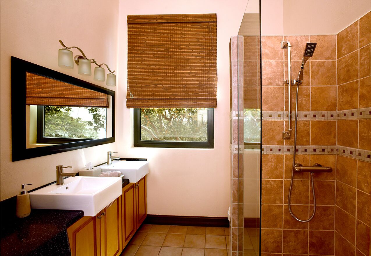 Une salle de bain avec deux lavabos, un miroir, une douche avec une porte en verre et une fenêtre avec un store.