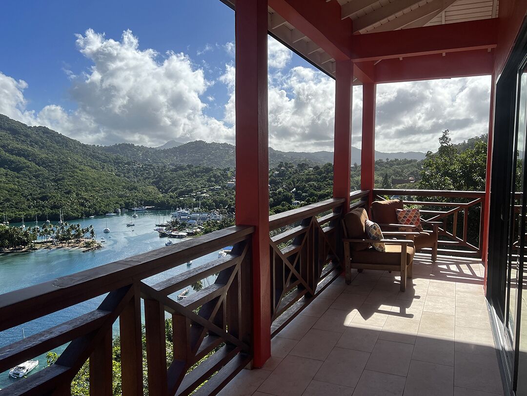 A balcony with a view of a river and mountains, wooden furniture, and a red railing.