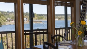 A dining area with a table set for two, overlooking a body of water with a dock and buildings in the distance. - South Whidbey with water view, beach and hiking  (Clinton)