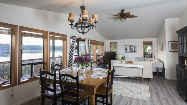 A dining area with a wooden table, chairs, and a chandelier. There are large windows with a view of a body of water. - South Whidbey with water view, beach and hiking  (Clinton)
