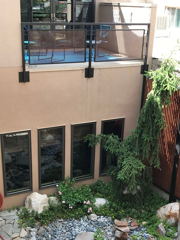 Un balcón con barandillas de malla, un pequeño jardín con rocas y flores, y vistas a un edificio con múltiples ventanas.