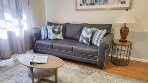 A living room with a grey sofa, floral cushions, a round wooden coffee table, a small side table with a lamp, and a framed picture on the wall.