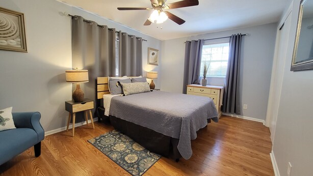 A bedroom with a bed, two bedside tables, a ceiling fan, a window with curtains, and a blue armchair.