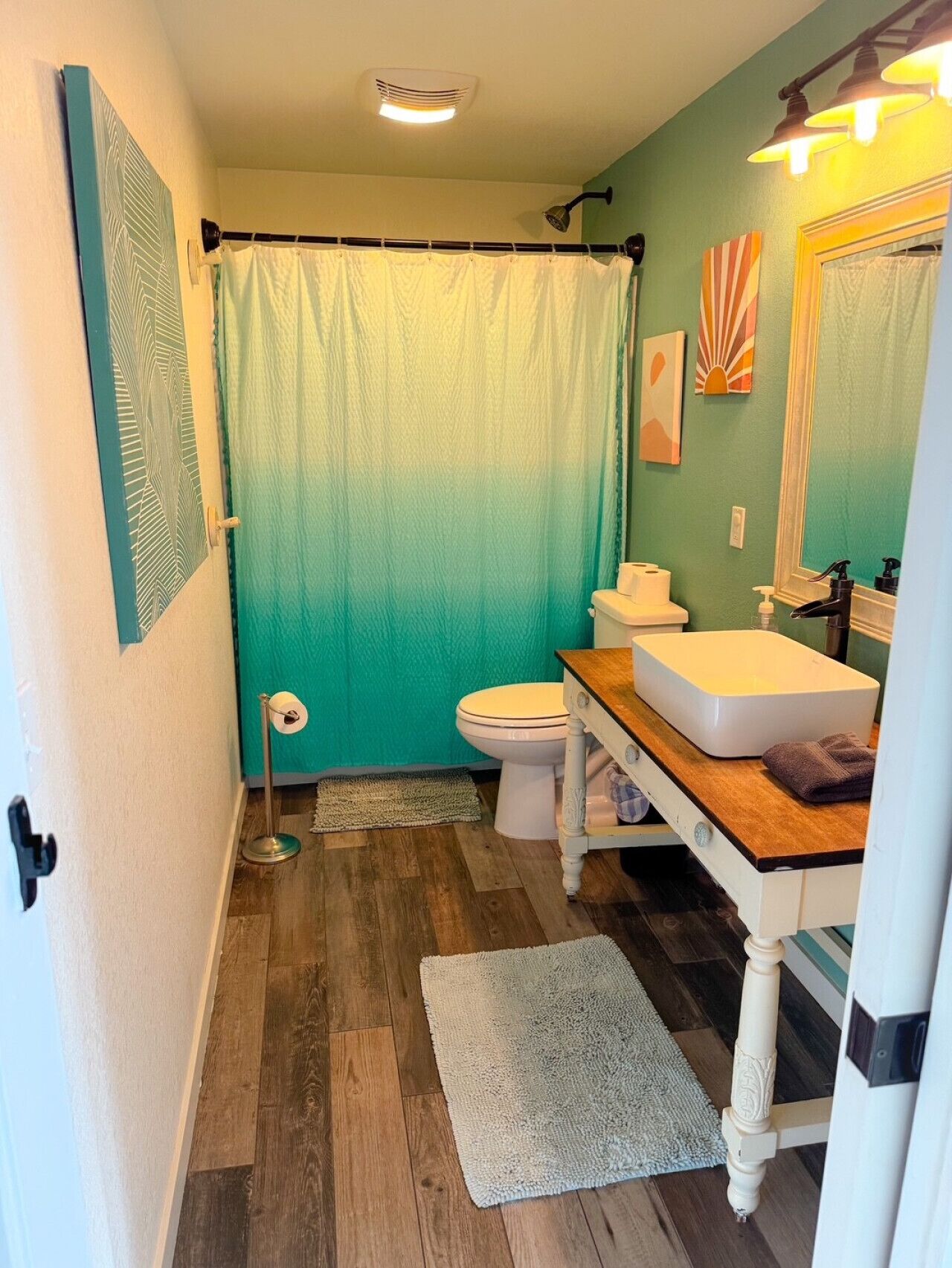 A bathroom with a white toilet, a wooden vanity with a white sink, a green shower curtain, and a mirror above the sink.