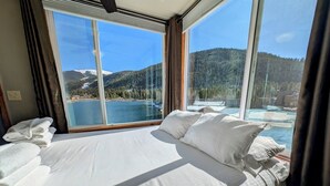 A hotel room with a large bed, white linens, and a view of a lake and mountains through large windows.