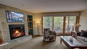 A living room with a stone fireplace, a television mounted on the wall, a patterned armchair, a sofa, and a balcony with outdoor furniture. - Family-Friendly Escape | Balcony & Scenic Views (Keystone)