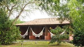 Un bâtiment rustique avec un toit en tuiles, entouré d’une végétation luxuriante et agrémenté d’un hamac tendu entre deux arbres.
