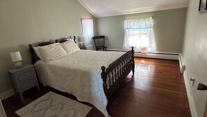 A bedroom with a large bed, a nightstand, a chair, a window with curtains, and a wooden floor. - Heart of the Seneca Lake Wine Trail (Burdett)