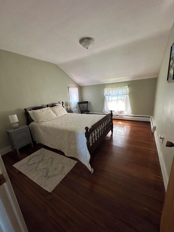 Un dormitorio con una cama grande, una mesita de noche, una silla, un ventanal con cortinas y suelo de madera.
