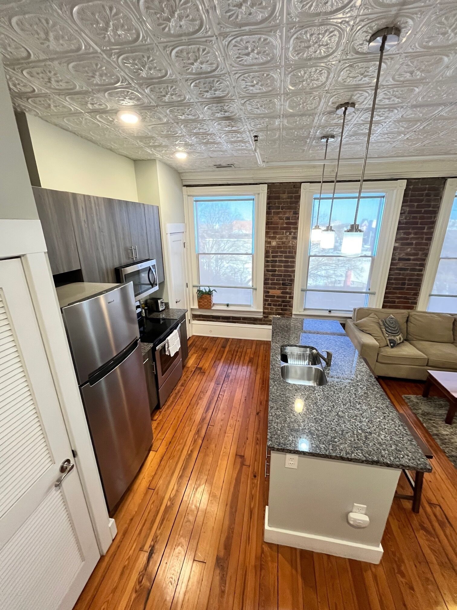 A modern kitchen with a stainless steel refrigerator, a built-in oven, and a granite countertop. There is a large window providing natural light and a view of the outdoors.