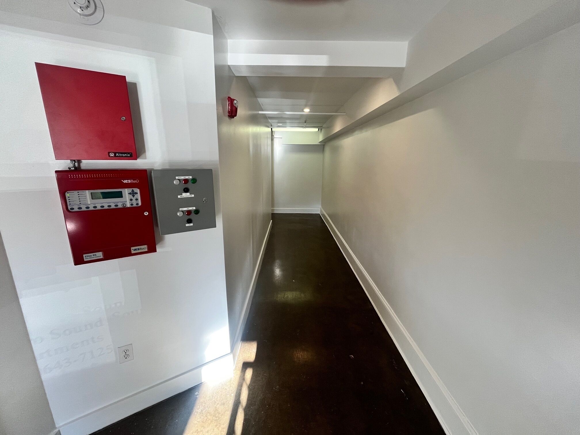 Un couloir avec un détecteur d’incendie rouge et un panneau de commande au mur.