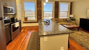 Una cocina moderna con encimera de granito, electrodomésticos de acero inoxidable y microondas integrado. Cuenta con un amplio ventanal que proporciona luz natural y vistas al exterior.