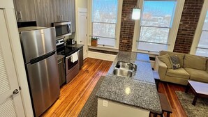 Una cocina moderna con horno empotrado, microondas y refrigerador de acero inoxidable. Hay una pequeña isla con pileta y un área de comedor con sofá y mesa de centro. El ambiente tiene pisos de madera y un techo decorativo.