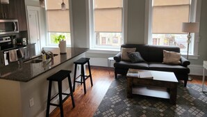 A modern kitchen with a central island, bar stools, a black sofa with patterned cushions, and a view of the street through large windows.