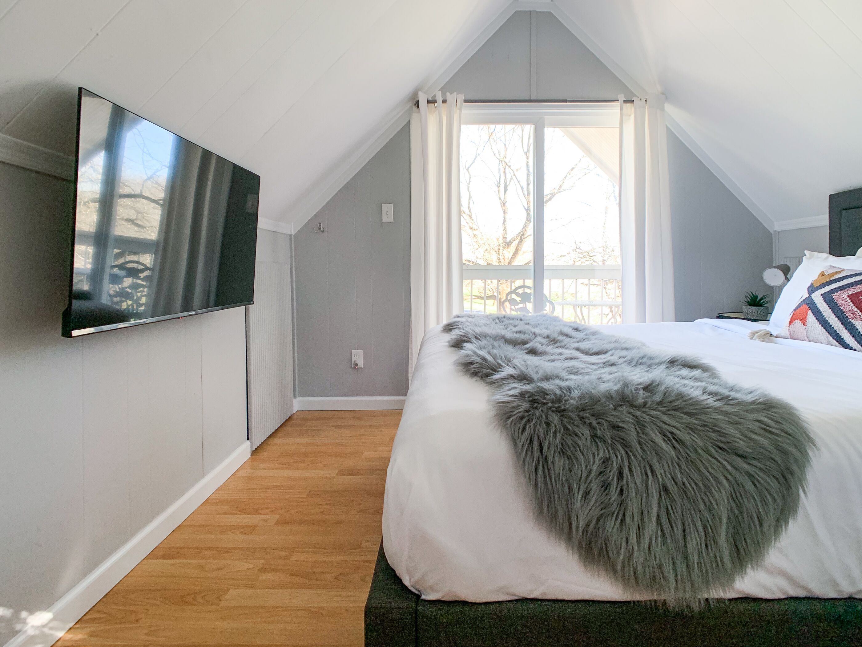 Une chambre à coucher avec un lit, un téléviseur fixé au mur, une fenêtre avec des rideaux et une vue sur l’extérieur.