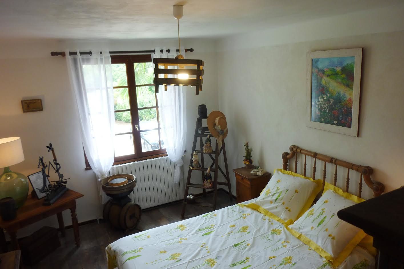 Una stanza con un letto, una finestra con tende, una scala di legno, un tavolino con una lampada e un quadro appeso al muro.