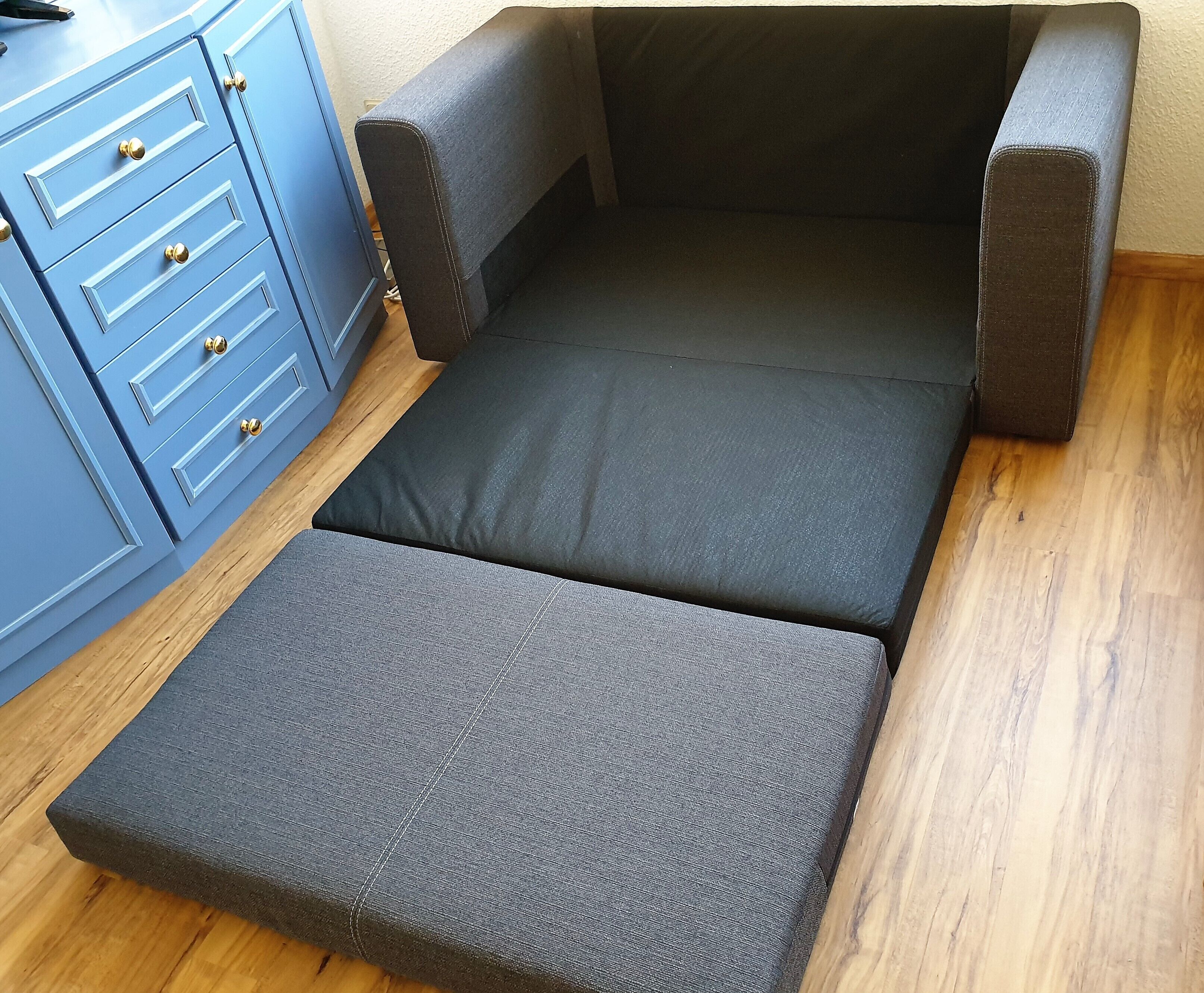 A small, compact bed with a grey mattress and matching headboard and footboard, positioned next to a blue dresser with multiple drawers and gold knobs.
