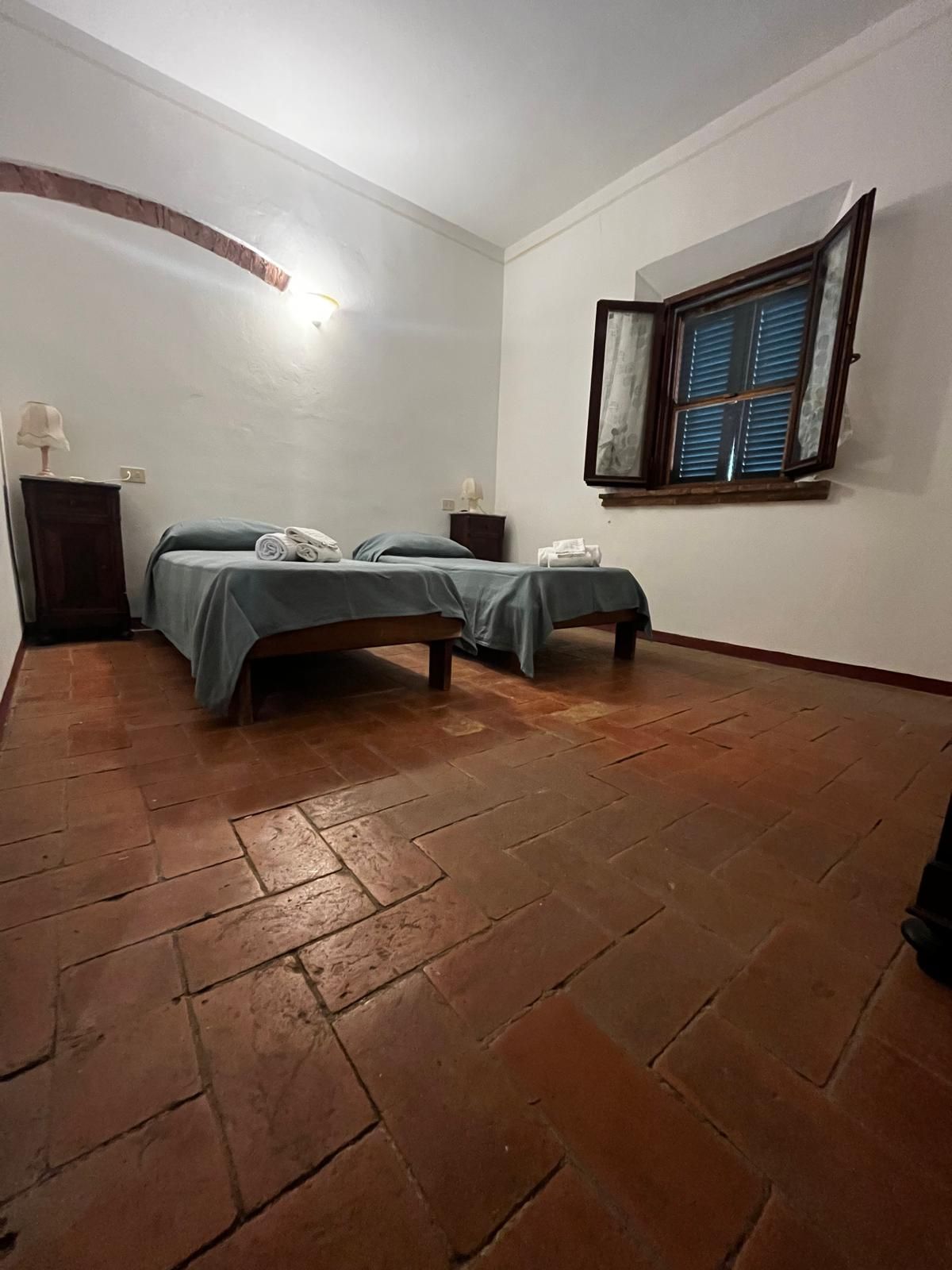A room with two beds, a window with blinds, a lamp on a nightstand, and a tiled floor.