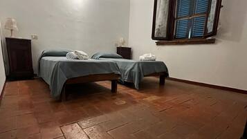 A room with two beds, a window with blinds, a lamp on a nightstand, and a tiled floor.
