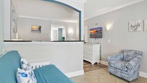 A living room with a sofa, a patterned armchair, a kitchen area with a counter, and a dining area with a table and chairs.