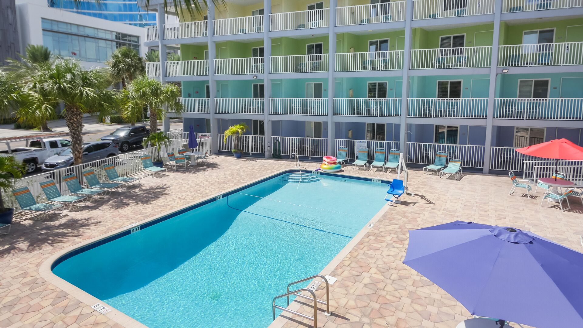 A swimming pool with lounge chairs and umbrellas, surrounded by a multi-story building with balconies.