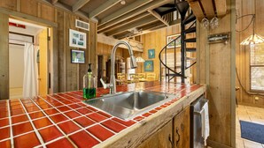 A kitchen with a red-tiled countertop, a stainless steel sink, wooden cabinets, and a staircase leading to an upper level. - Hook, Wine & Sinker Cottage: Water Views, Kayaks, Bikes & Old Florida Charm (Cedar Key)