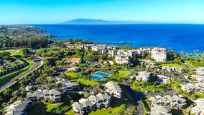 Vue aérienne d’un quartier résidentiel situé sur la côte, avec un grand complexe hôtelier, un court de tennis, et une vue sur l’océan et une montagne au loin.