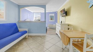 A room with a blue sofa, a wooden table with chairs, a TV mounted on the wall, and a window with blinds.