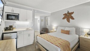 A compact hotel room with a kitchenette, a bed with a patterned blanket, and a wall-mounted decorative piece.