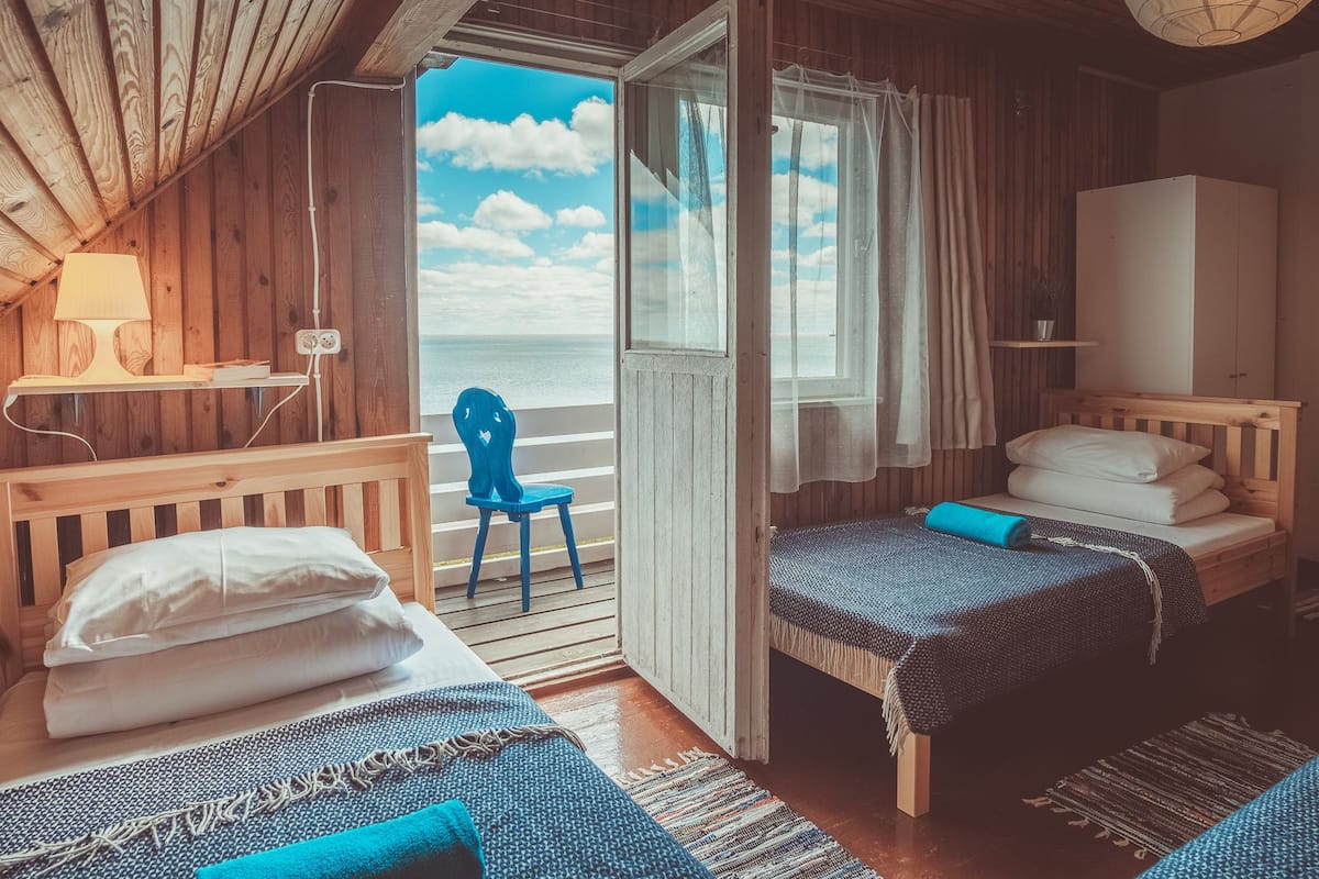 A cozy room with wooden walls and a large window overlooking the sea. There are two beds, a chair, and a desk with a lamp.