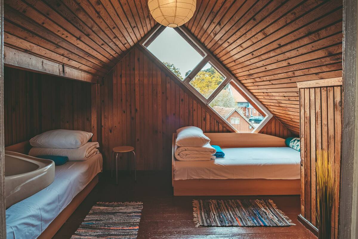 A cozy attic bedroom with wooden walls, a skylight, two beds, and a small stool.