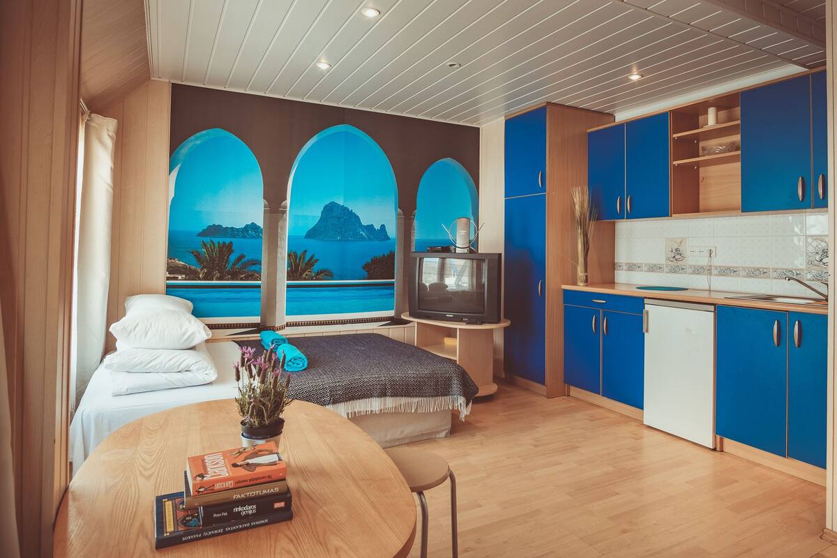 A modern room with a bed, a TV, a dining table with books, and a view of the sea through arched windows.