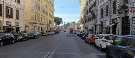 Katu, jonka varrella on parkissa autoja ja jonka molemmin puolin on rakennuksia, joissa on erilaisia kylttejä, kuten esimerkiksi kyltti, jossa lukee ”Pizzeria”, ja kyltti, jossa lukee ”Baari”.