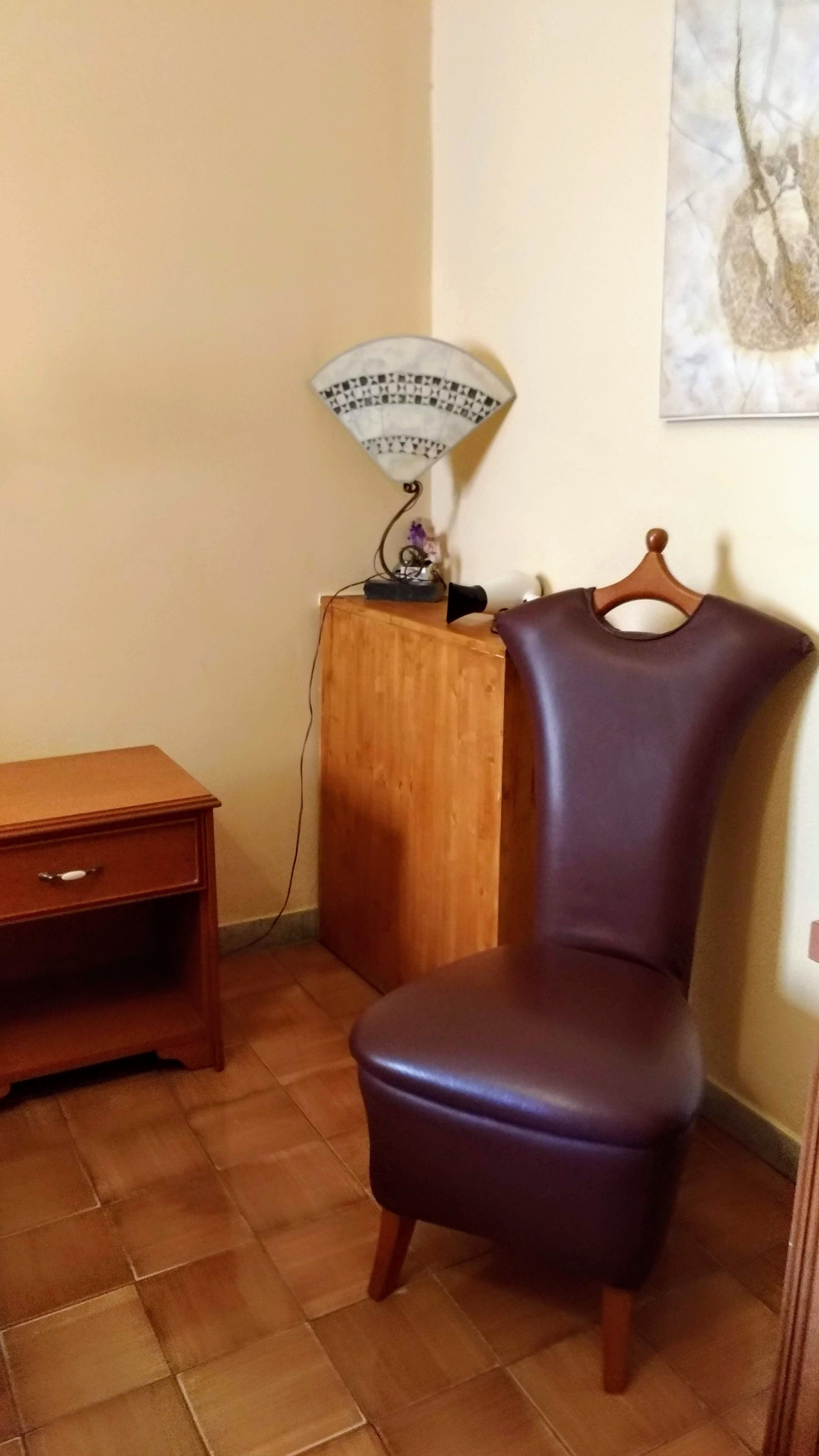 A corner room with a wooden nightstand, a purple chair, a wooden cabinet, and a wall-mounted lamp.