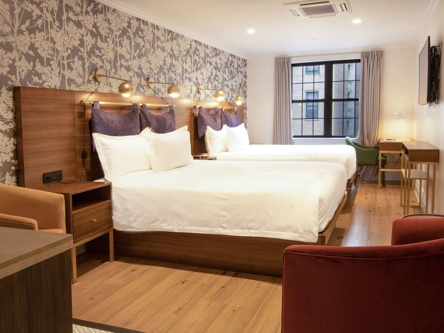 A hotel room with two beds, a desk, a chair, and a window with a view of the city.