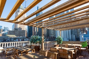 Een terras met houten tafels en stoelen, plantenbakken en uitzicht op de skyline van de stad.
