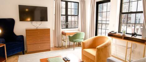 A modern living room with a sofa, armchair, wooden coffee table, and a desk with a chair. A TV is mounted on the wall, and there are large windows with curtains.