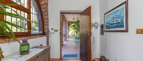 Un couloir avec un banc en bois, une commode décorative, une porte donnant sur un jardin et un tableau encadré accroché au mur.
