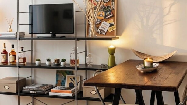 A modern living room with a wooden dining table, a TV on a metal stand, a shelf with books and decorative items, and a small plant.