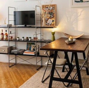 Una sala de estar moderna con una mesa de comedor de madera, un televisor sobre un soporte metálico, una estantería con libros y objetos decorativos, y una pequeña planta.