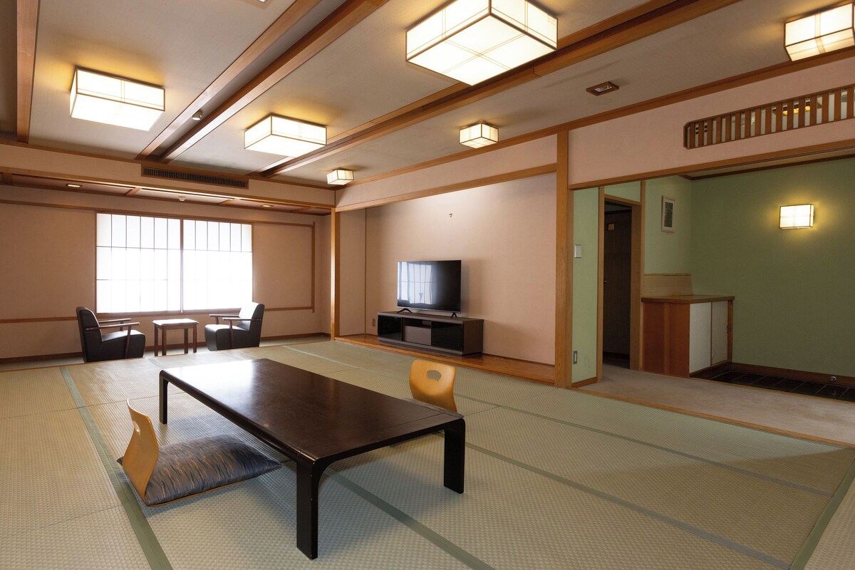 A traditional Japanese-style room with tatami flooring, a low table, chairs, a television, and sliding doors.