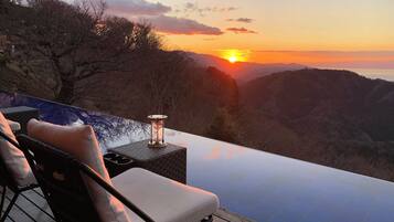 A sunset view from an infinity pool with a chair and a table, overlooking a mountainous landscape.